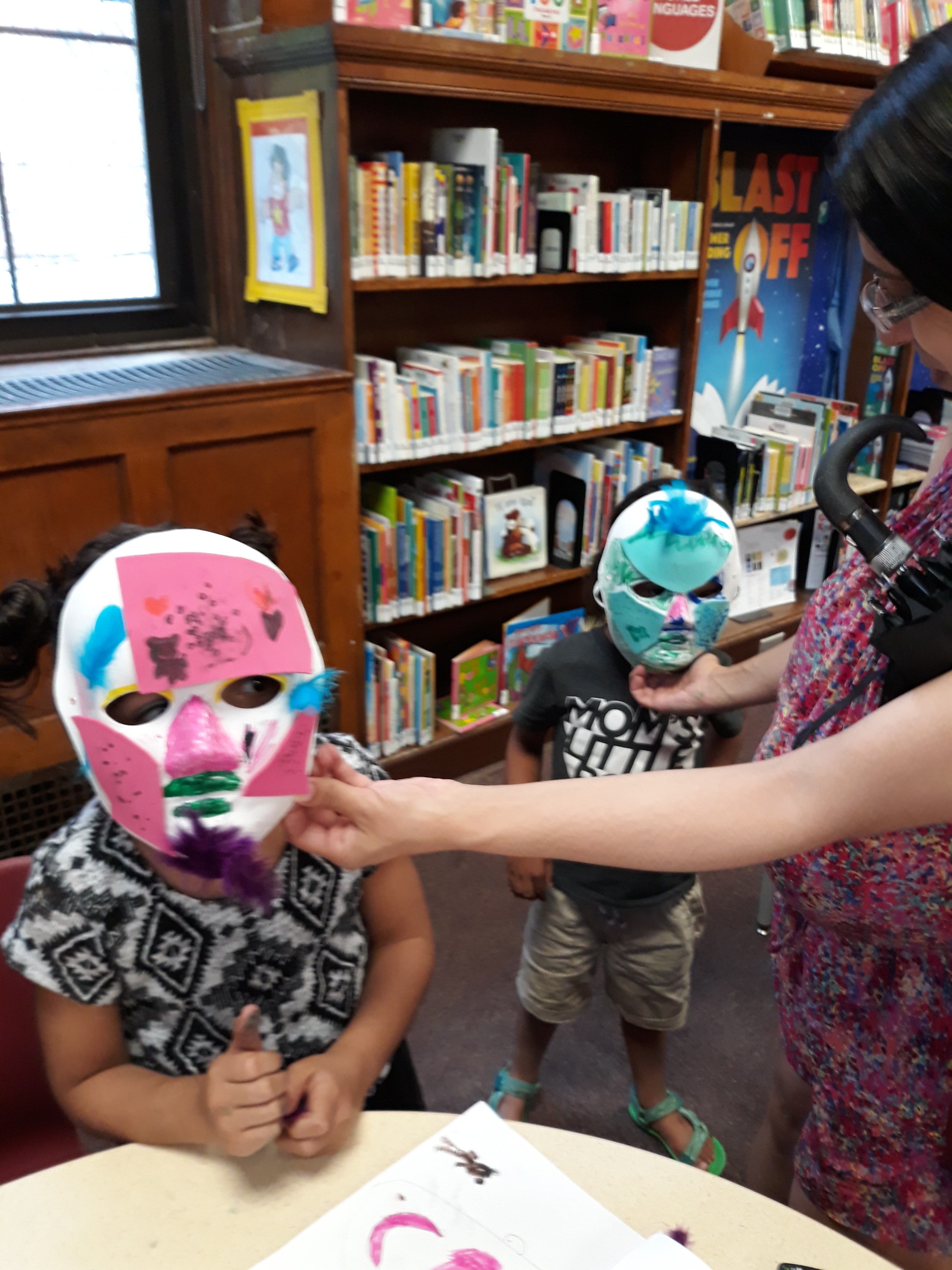 Two library-patrons-turned-imaginary-creatures get help from an adult in showing off their colorful masks.